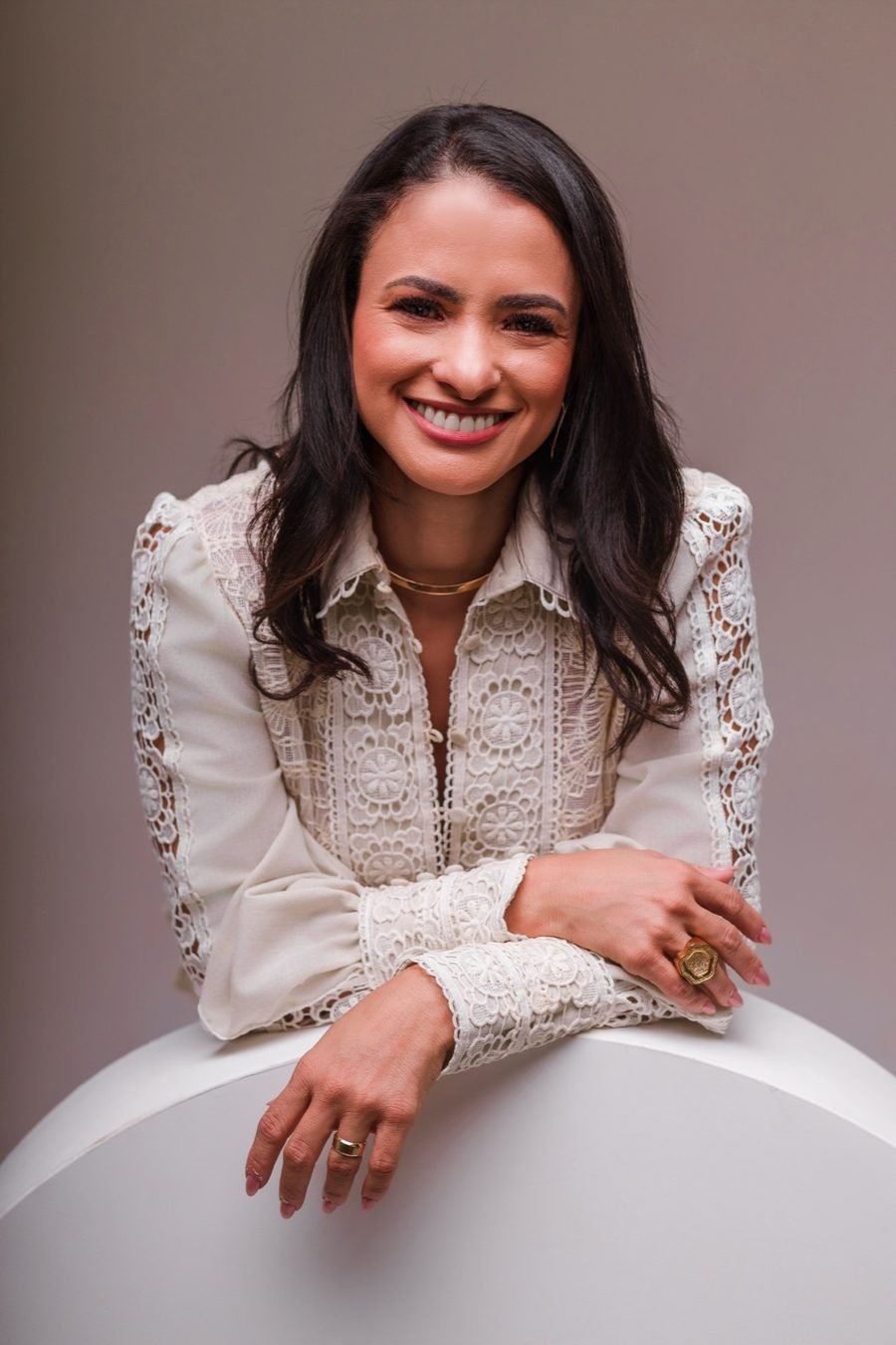 Kleiciane Rocha sorrindo, de camisa de renda clara, sobre fundo rosé